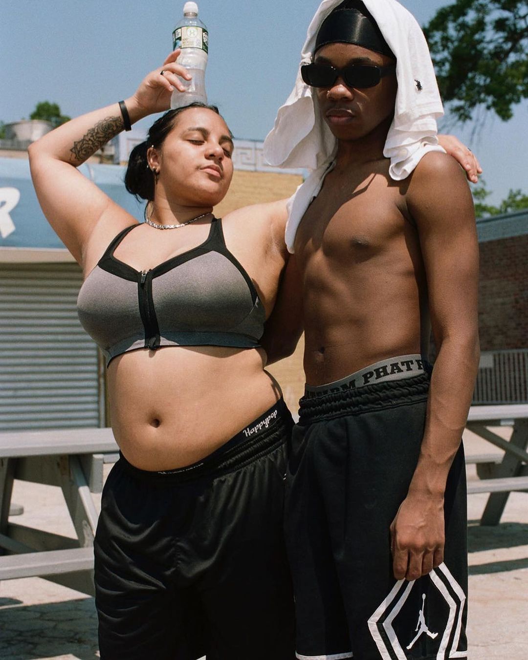 Two young adults pose outdoors on a sunny day. The woman in a sports bra balances a water bottle on her head.