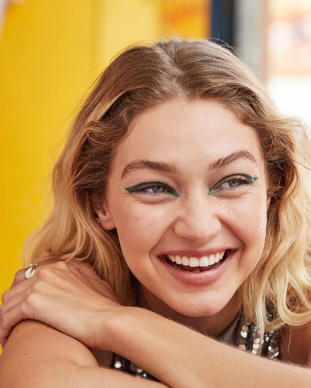 Close-up of a smiling woman with blond hair and green eyeliner, posing in front of a yellow background, capturing a joyful expression.