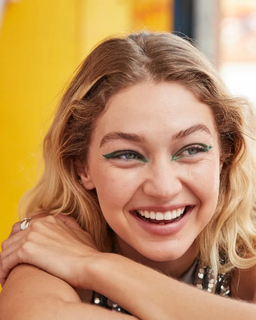 Close-up of a smiling woman with blond hair and green eyeliner, posing in front of a yellow background, capturing a joyful expression.