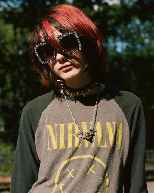 Teen with red-streaked hair wears jeweled sunglasses and Nirvana T-shirt outdoors among green trees in a fashion portrait.