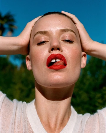 Portrait of a woman with red lipstick and slicked-back hair, eyes closed, hands on head, outdoors with a clear sky and green trees.