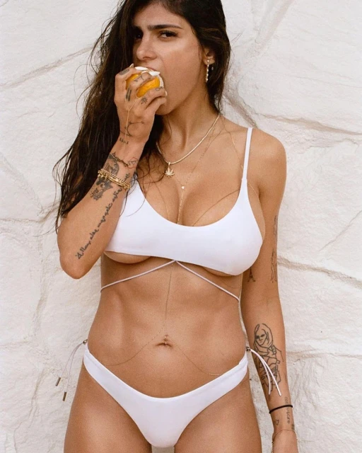 Adult woman in a white bikini eating a piece of citrus fruit, standing in front of a textured white wall in a medium shot.