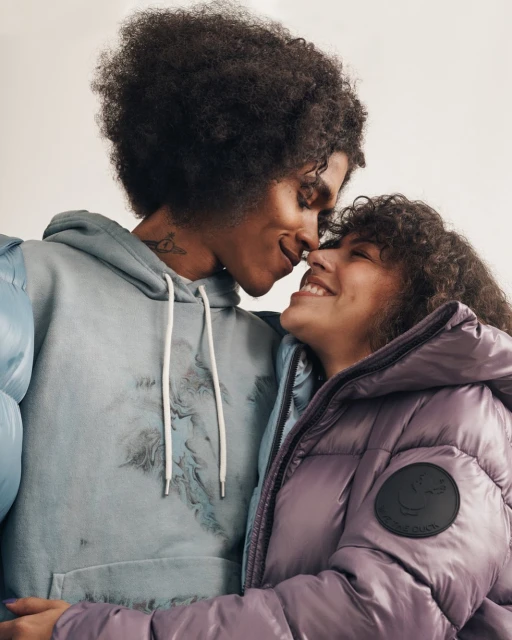 A happy couple in a close-up indoor shot. The man is wearing a hoodie and the woman a lavender puffer jacket, both smiling.