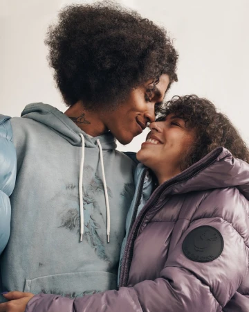 A happy couple in a close-up indoor shot. The man is wearing a hoodie and the woman a lavender puffer jacket, both smiling.