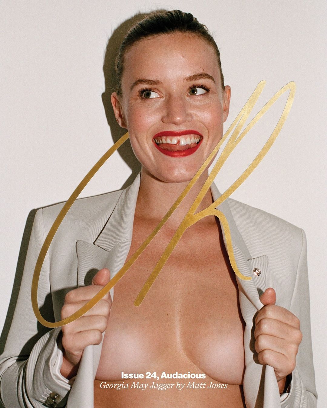 Smiling model Georgia May Jagger poses in a fashion editorial, opening her blazer to expose her chest, with a large gold text overlay.