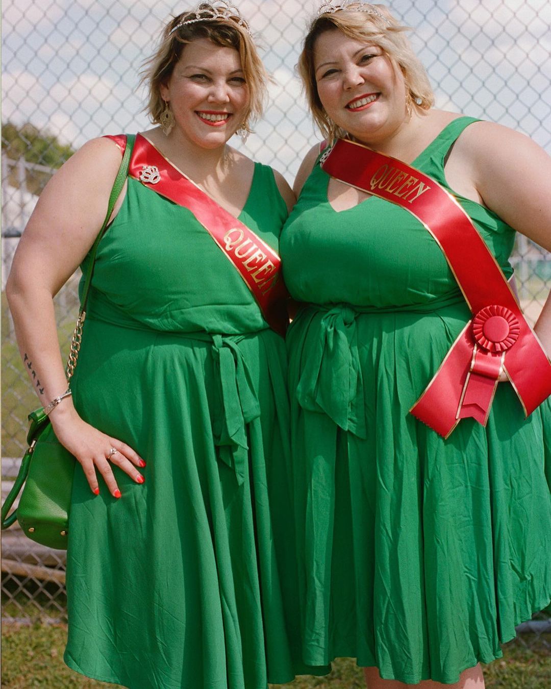 Two smiling women in green dresses with red sashes labeled 'Queen' stand outdoors, wearing tiaras. The background is blurred.