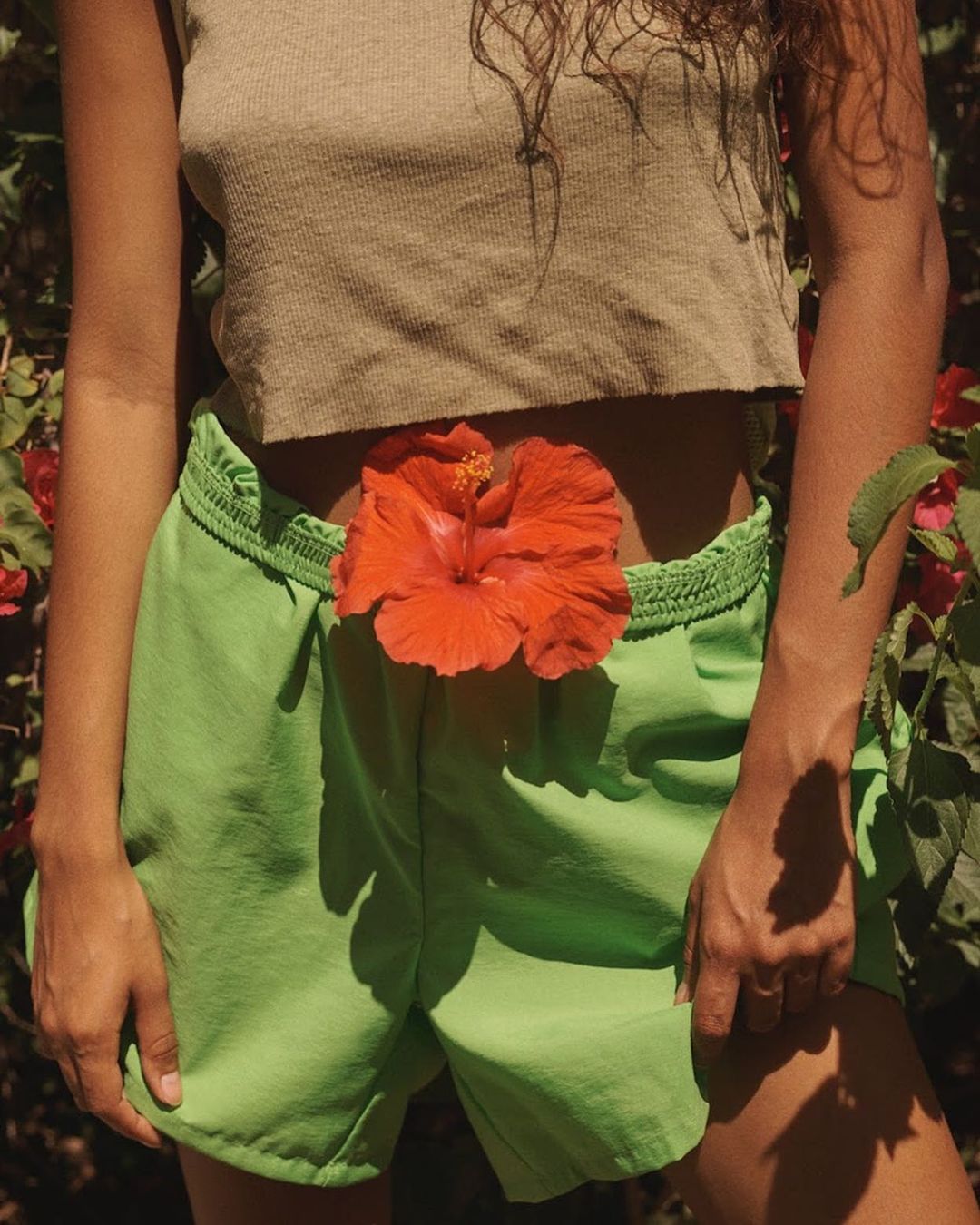 Waist-up shot of a person wearing green shorts and a beige top with a large orange hibiscus flower placed at the waist.