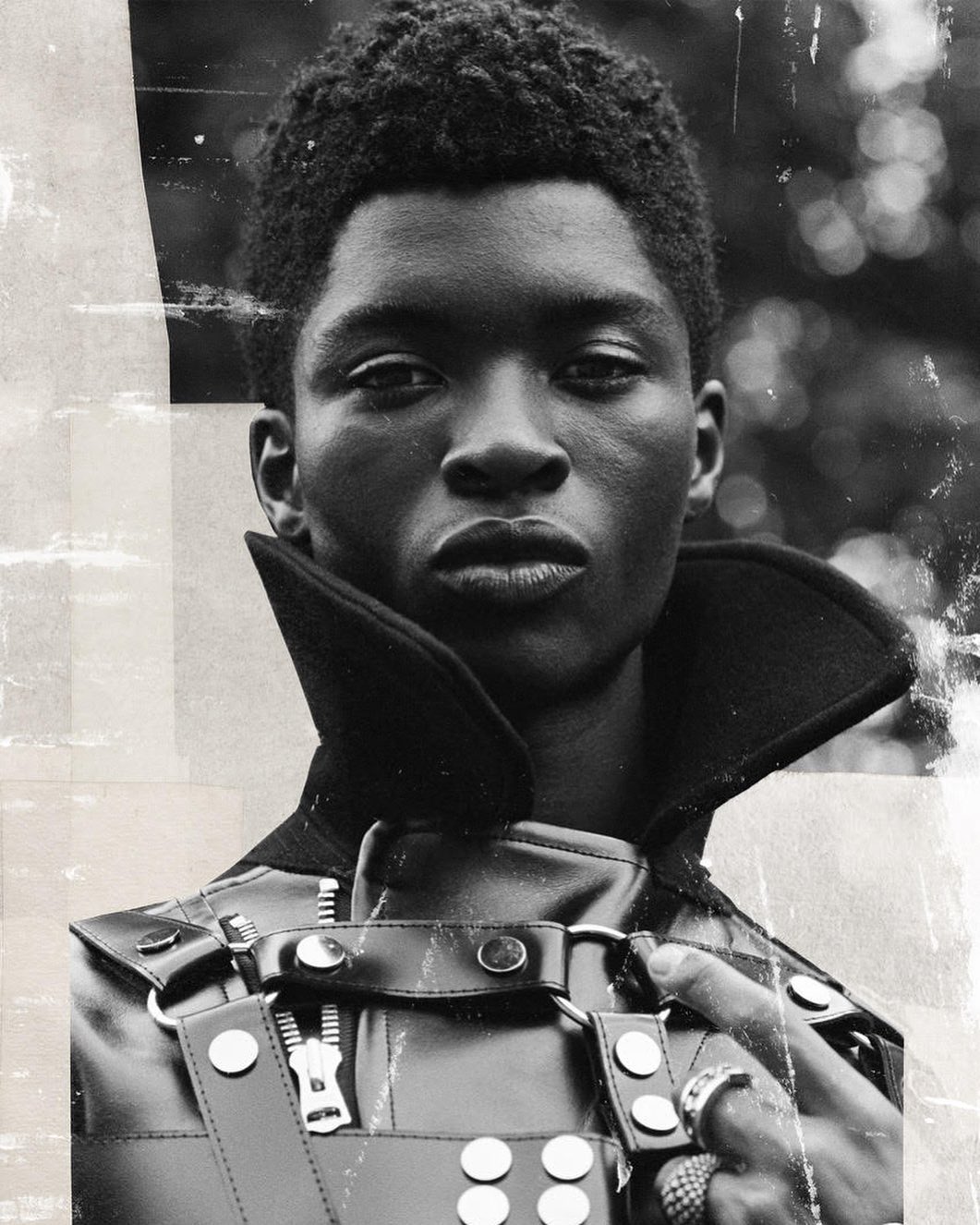 Monochrome portrait of a Black male model wearing a structured leather outfit with a high collar against a blurred background.