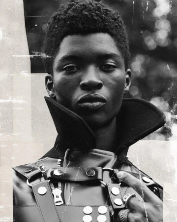Monochrome portrait of a Black male model wearing a structured leather outfit with a high collar against a blurred background.