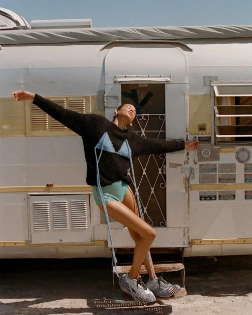 Woman posing in a blue crochet bikini top and teal shorts next to a silver trailer on a sunny day, arms outstretched.