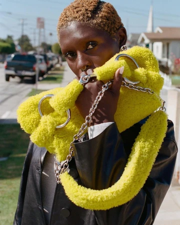 A Black woman poses outdoors with a bright yellow, furry handbag with a silver chain strap and rings, wearing a black leather jacket.