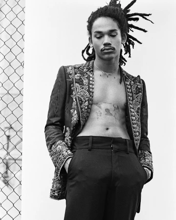 Black and white portrait of a man with dreadlocks wearing an embroidered blazer and dark trousers.