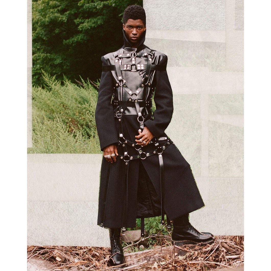 A striking portrait features a Black male fashion model in a dark coat and leather harness, posing outdoors against a natural backdrop.