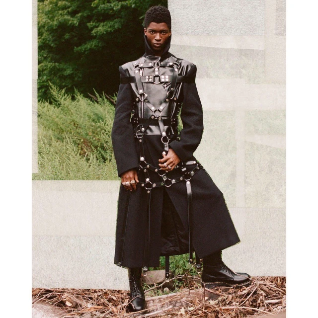 A striking portrait features a Black male fashion model in a dark coat and leather harness, posing outdoors against a natural backdrop.