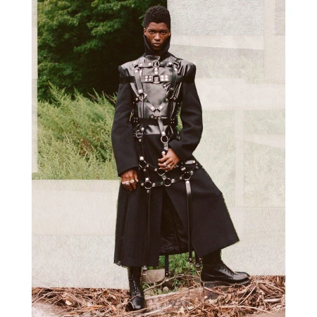 A striking portrait features a Black male fashion model in a dark coat and leather harness, posing outdoors against a natural backdrop.