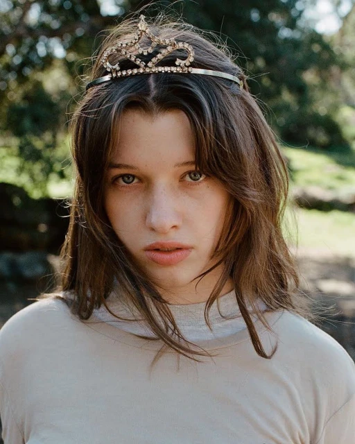 Portrait of a young adult female with dark hair, wearing a tiara, gazing directly at the camera in a natural outdoor setting.