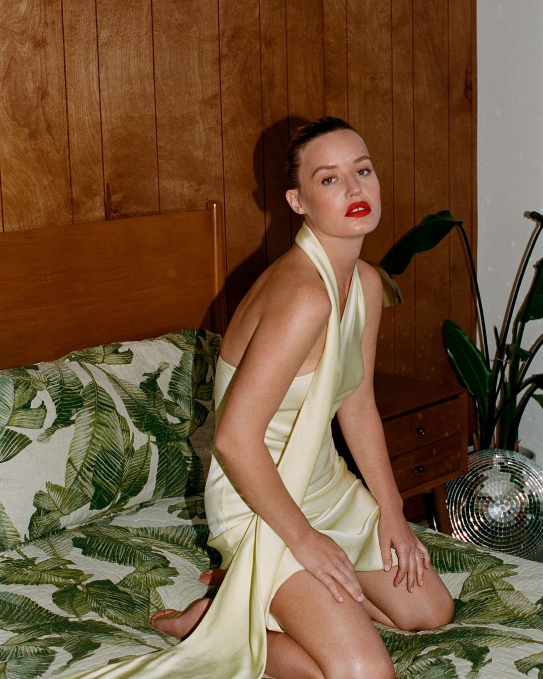 Woman in pale yellow dress sits on bed with tropical print, wood paneling, houseplant, and disco ball in the background.