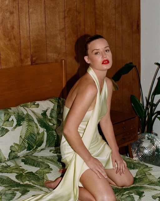 Woman in pale yellow dress sits on bed with tropical print, wood paneling, houseplant, and disco ball in the background.