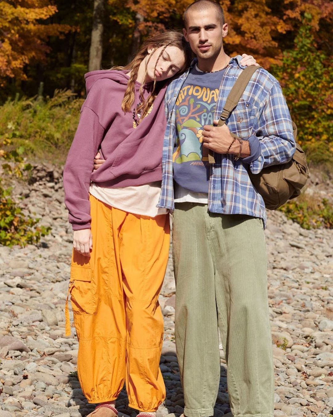 A stylish couple poses outdoors among autumn trees; she wears a plum hoodie with orange pants, and he wears a plaid shirt and green pants.