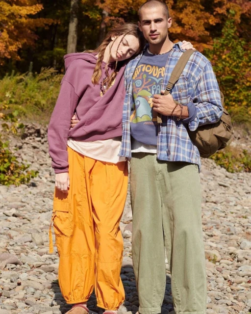 A stylish couple poses outdoors among autumn trees; she wears a plum hoodie with orange pants, and he wears a plaid shirt and green pants.