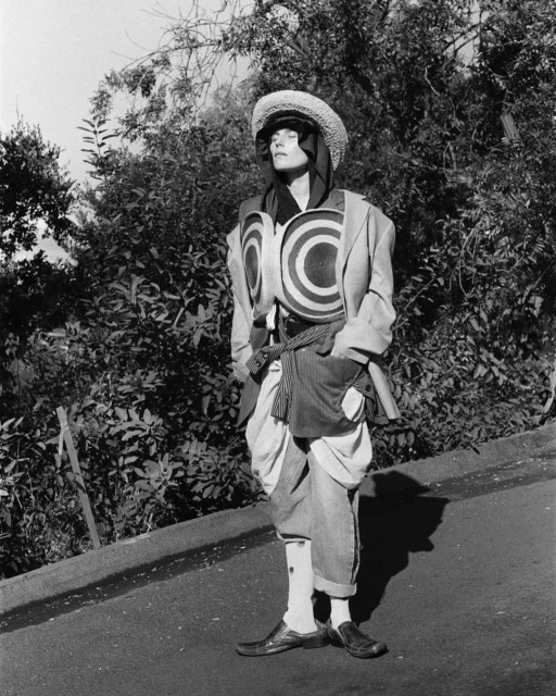 Monochrome full shot of a person wearing a straw hat, patterned vest, jacket, and pants outdoors surrounded by trees and greenery.