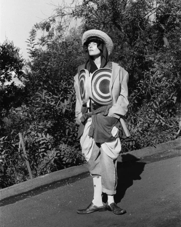 Monochrome full shot of a person wearing a straw hat, patterned vest, jacket, and pants outdoors surrounded by trees and greenery.