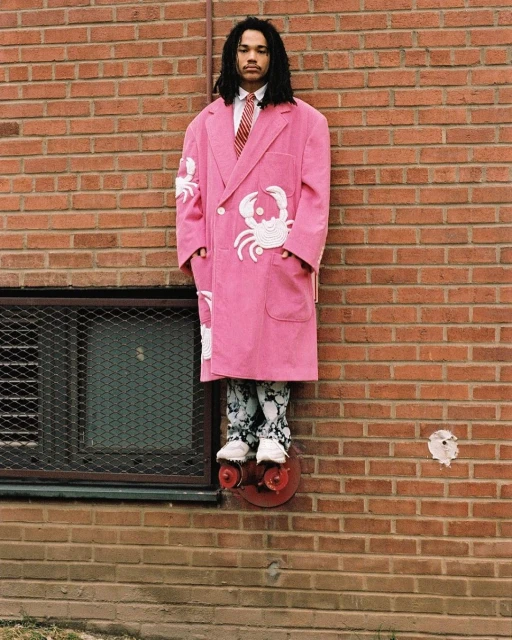 Young adult in pink crab coat, patterned pants, stands on outdoor fire hydrant. Brick building backdrop, urban fashion statement.
