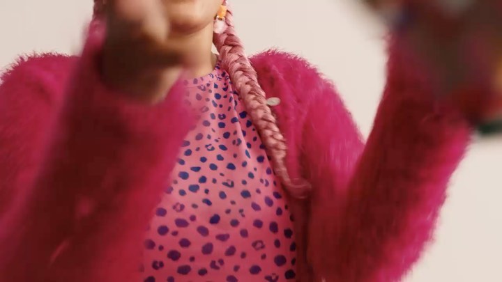 A woman with pink braided hair wears a pink fuzzy jacket over a pink patterned shirt, set against a white background.