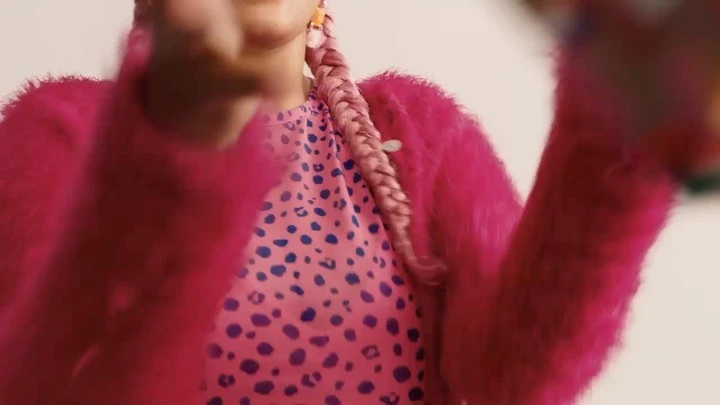 A woman with pink braided hair wears a pink fuzzy jacket over a pink patterned shirt, set against a white background.
