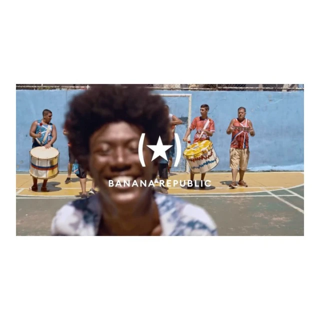 An out-of-focus man smiles in a close-up shot. Behind him, out of focus, are 3 men with drums on an outdoor sports court.