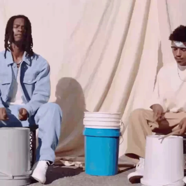 Two young men play bucket drums outdoors in soft light; a unique musical performance and display of rhythm.