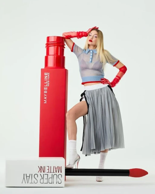 A fair-skinned woman with long blonde hair poses with red lipstick, a grey pleated skirt, red gloves, and a sheer patterned top in a studio.