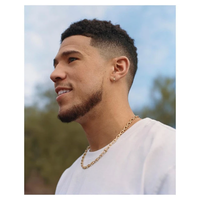 Portrait of a smiling adult male with a short haircut and beard, wearing a white t-shirt and gold chain against a cloudy sky backdrop.