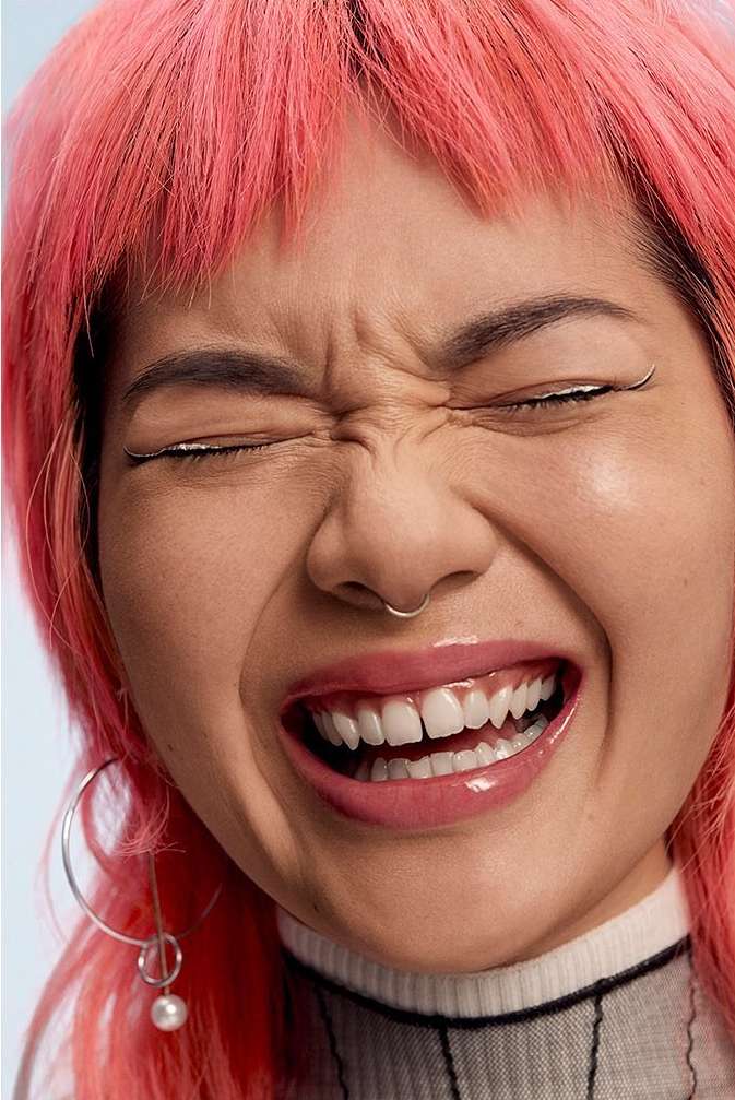 Close-up portrait of a young adult woman with pink hair laughing with her eyes closed, wearing hoop earrings and a white and black top.