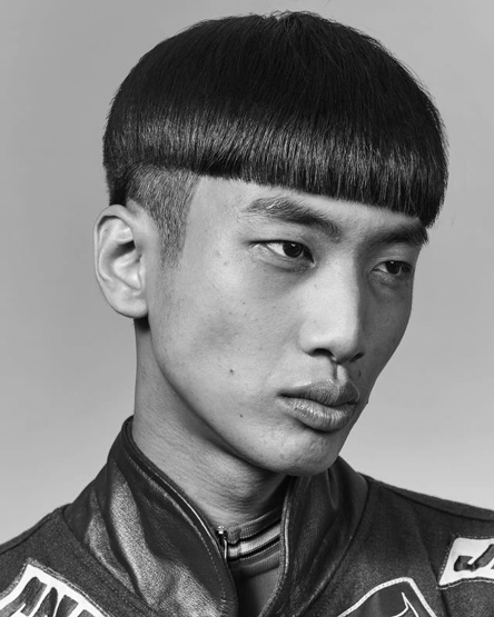 Portrait of Asian man with bowl cut hairstyle wearing a black leather jacket against a neutral gray studio backdrop in black and white.