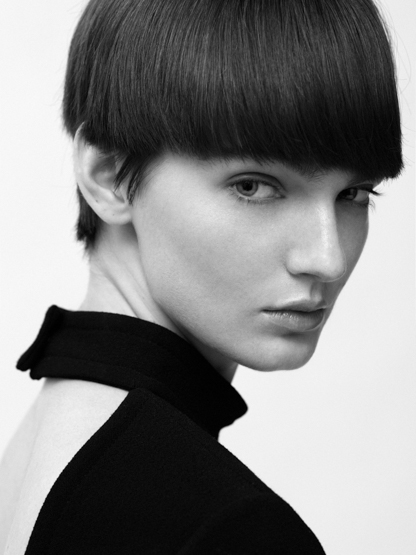 Headshot of a fair-skinned adult woman with a dark bowl cut wearing a black top against a white background in a portrait.