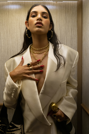 Brunette woman in white blazer with layers of gold necklaces and rings poses indoors with direct eye contact and red nail polish.
