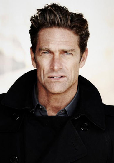 Headshot of handsome adult male with light eyes and dark hair wearing a dark coat and gray shirt against a neutral backdrop.