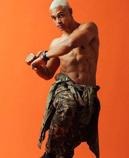 Shirtless man with bleached hair poses against an orange background, wearing army-style pants tied around his waist and a smart watch.