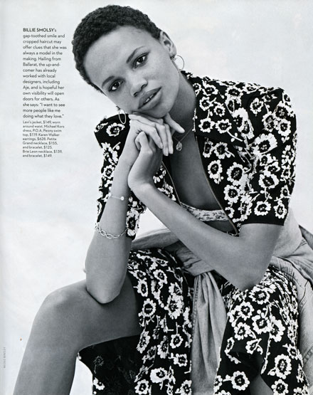 Black-and-white fashion shot of a female model with short hair wearing a floral dress and jacket, seated with hands near her face.