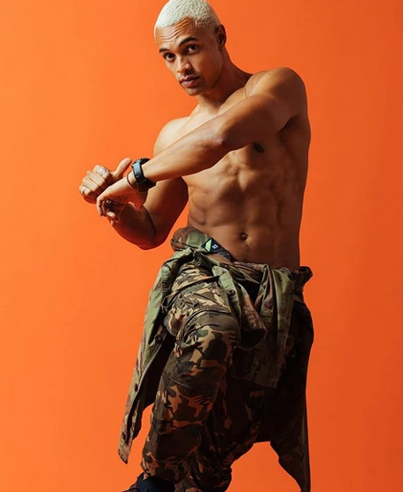 Shirtless male model with blonde hair wearing camouflage cargo pants posing against a vibrant orange backdrop.