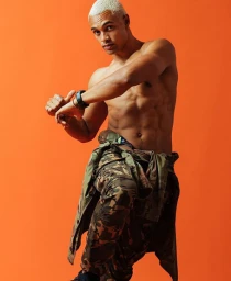 Shirtless male model with blonde hair wearing camouflage cargo pants posing against a vibrant orange backdrop.