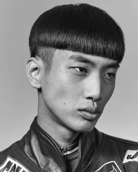 Monochrome portrait of an Asian male model with a bowl cut hairstyle wearing a dark patterned jacket against a neutral studio background