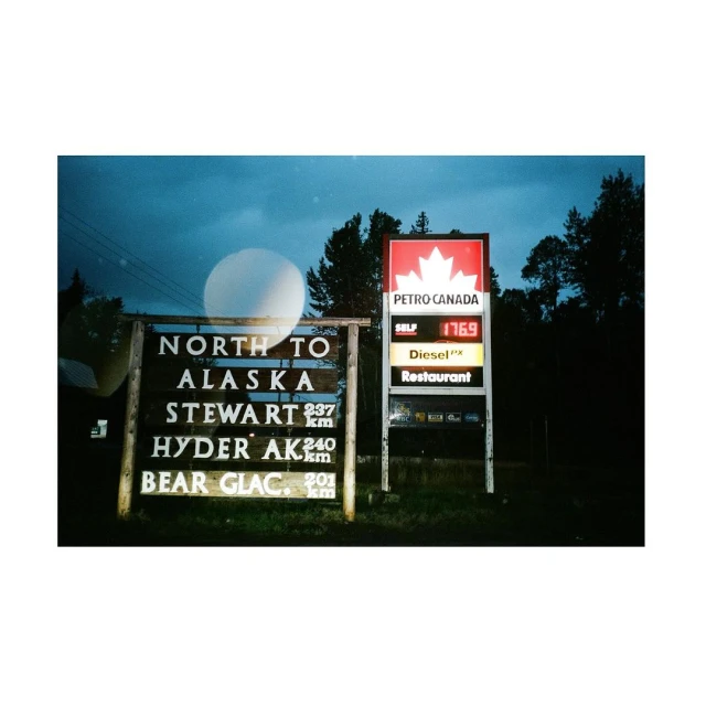 Roadside sign for Petro Canada gas station with fuel prices and directions to Alaska, Stewart, and Hyder AK, surrounded by trees.