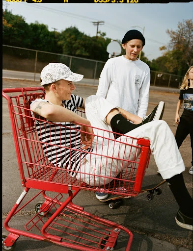 Three young adults pose outdoors. One sits in a red shopping cart, and two stand nearby, with skateboards visible on the ground.