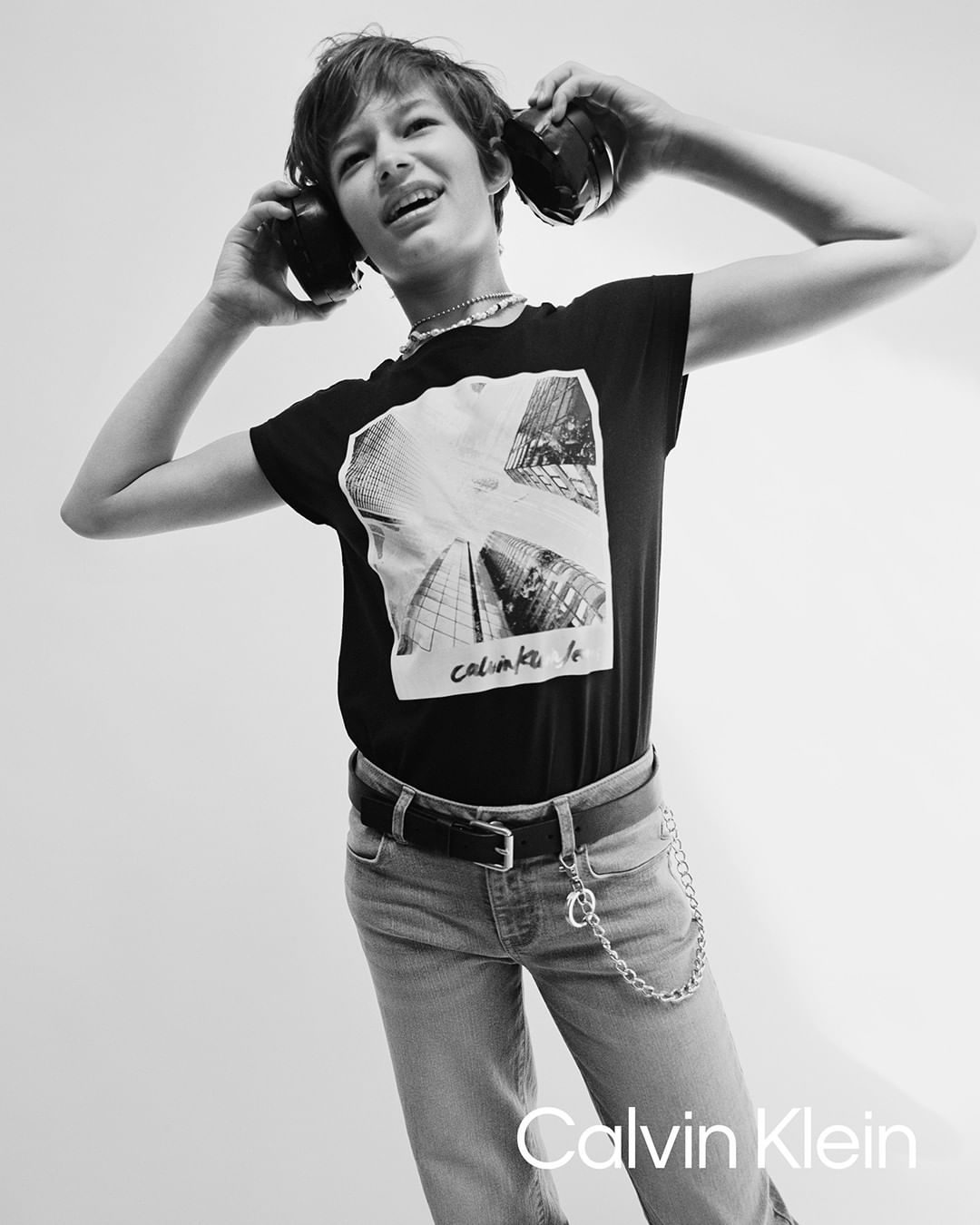 Teen poses in Calvin Klein advert wearing headphones, a graphic tee, jeans, and a chain belt against a stark white backdrop.