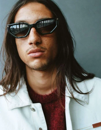 Close-up portrait of a man with long dark hair and freckles wearing geometric sunglasses and a white jacket by Matteo Montanari