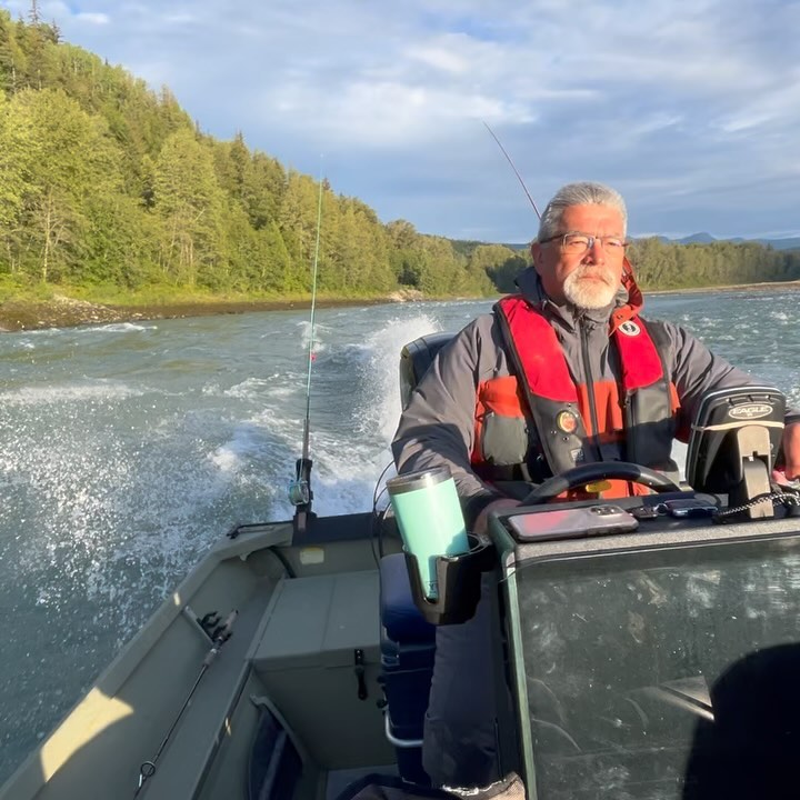 Older man navigates a river in a small motorboat wearing a life jacket, with forested hills and a cloudy sky in the background.