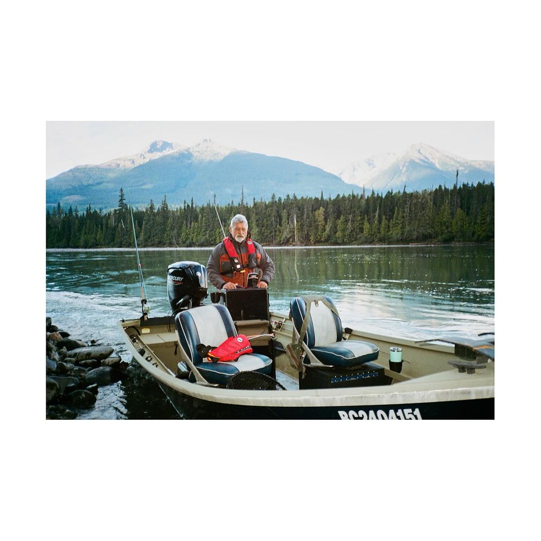 Elderly man wearing a life vest piloting a boat on a lake with mountains and trees in the background on a sunny day.