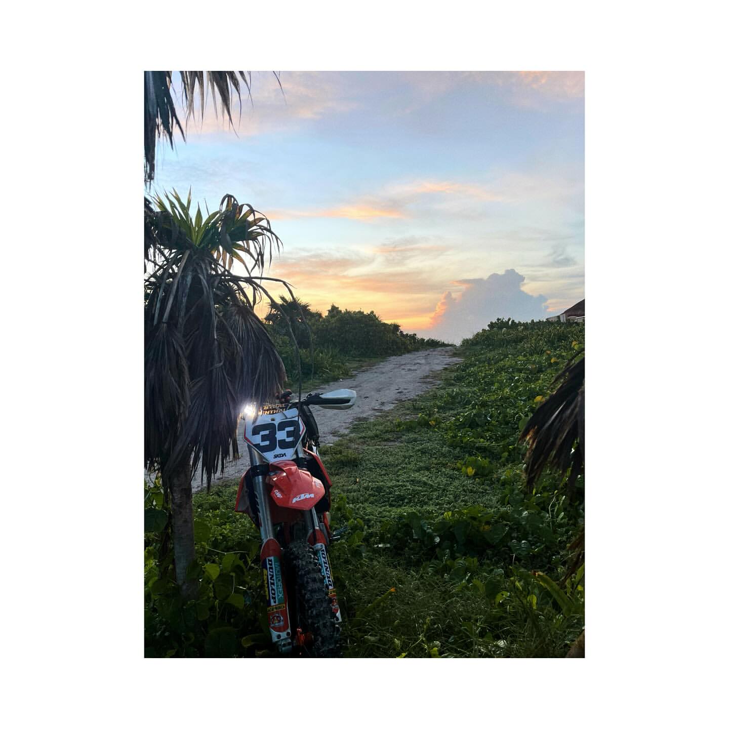 An off-road motorcycle is parked on a dirt path surrounded by green vegetation, with a vibrant sunset sky in the background.
